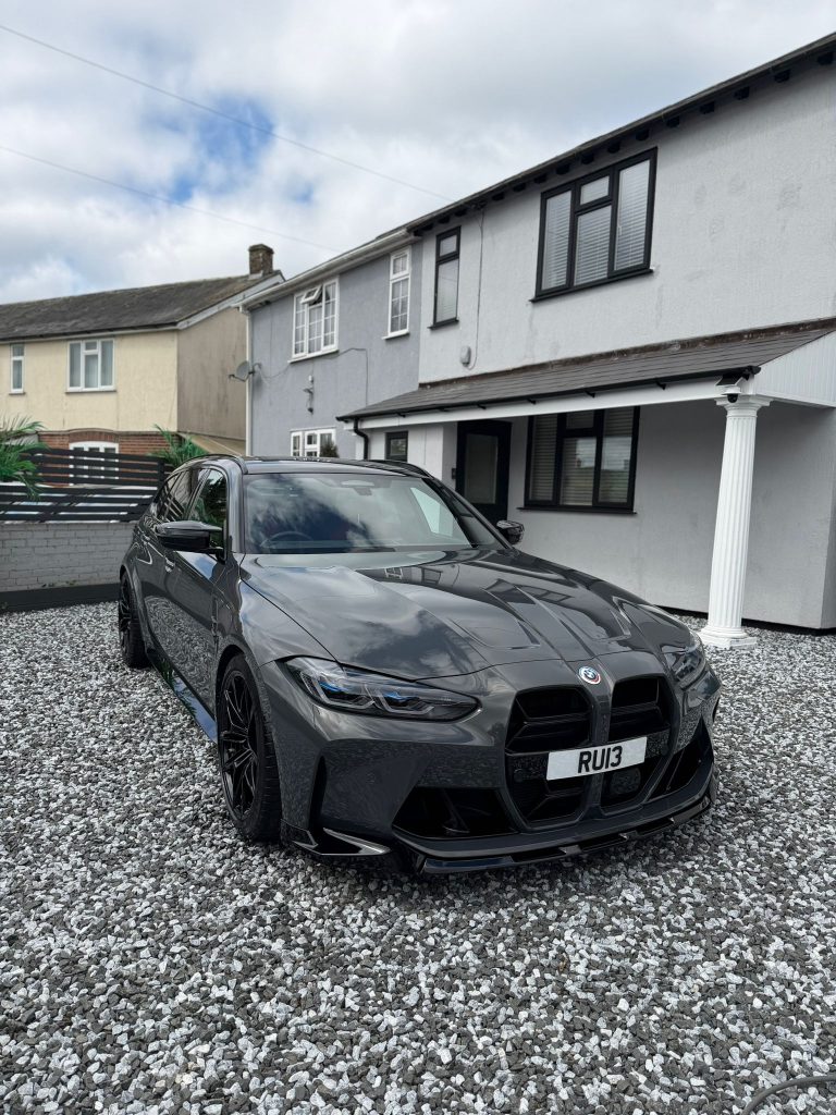 Grey BMW M3 Touring with gloss black wheels on a gravel drive after detailing